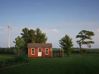 In a horizontal line from left to right, carefully spaced: a wind turbine, a red cabin with a tree behind it, and two more trees occupying similar space. The sky is blue and the lawn dark green; the evening light has already fallen off the lawn but is still hitting the arrayed objects.